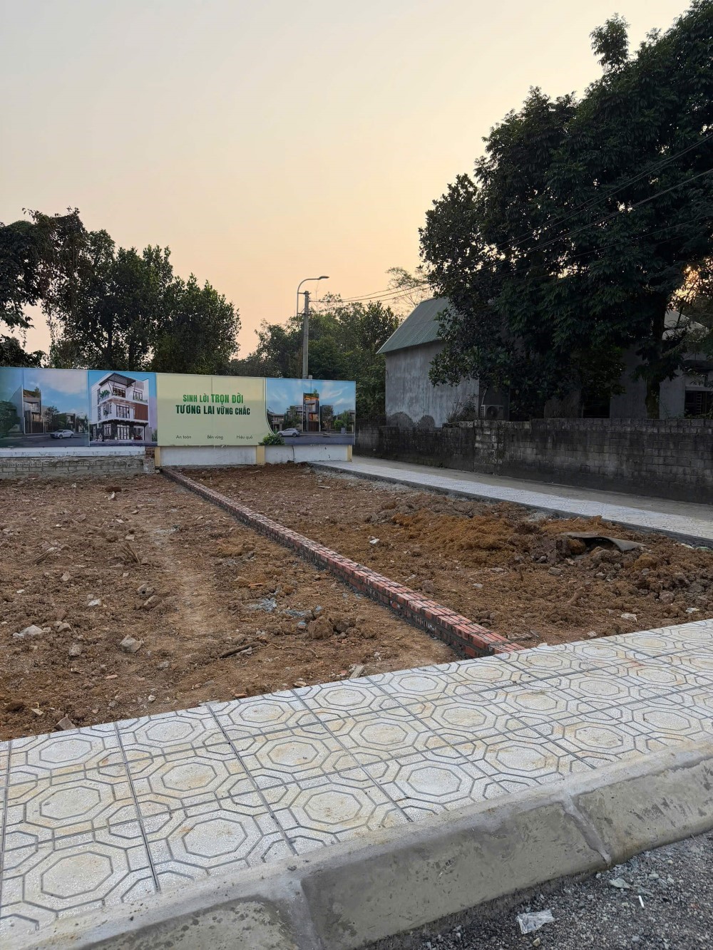 Lô đất nền đang san lấp, vỉa hè lát gạch, phía sau là bảng quảng cáo dự án nhà ở